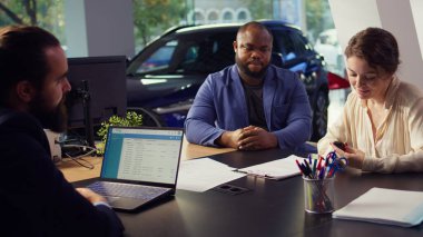 Deneme sürüşü bittikten sonra satış elemanı ve müşterilerin oturduğu ve konuştuğu bir araba galerisi görüntüsü. Ajan, araç kiralamak isteyen bir çiftle konuşuyor. Kamera B, kapat.