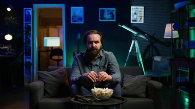 Tembel kanepe patatesi insanı düşüncesizce TV 'de programlar izliyor ve patlamış mısır yiyor, ekrana yakından bakıyor. Oturan adam abur cubur yerken akılsız eğlence tüketiyor, kamera B