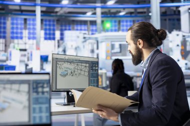 Güneş paneli fabrikasında müfettiş imalat ekipmanlarını kontrol ediyor, belge dosyalarını okuyor. Endüstriyel tesislerdeki adam yeşil enerji üretim sürecini inceliyor, evrak işlerini kontrol ediyor.