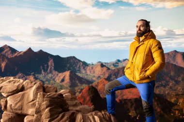 Sıcak kıyafetler giymiş yürüyüşçü zirveye tırmandıktan sonra bir dağın zirvesinde duruyor, engebeli tepelerin ve engin gökyüzünün nefes kesici manzarasına bakıyor. Manzaralı macera.