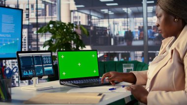 Genç genel müdür laptopta yeşil ekran kullanıyor ve personeli için performans ölçümlerini değerlendiriyor, bir planlama oturumu düzenliyor. Afrikalı Amerikalı CEO liderlik becerileri gösteriyor. Kamera B.