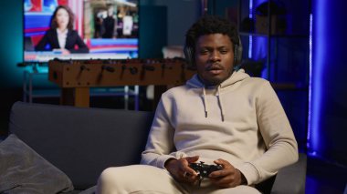 Afro-amerikalı oyuncu evde oturmuş koltuğa oturuyor, oyun oynamak ve boş zamanların tadını çıkarmak için heyecanlanıyor. Dekore edilmiş bir oturma odasında kanepede oturmuş, konsolda video oyunları oynuyor. Kamera A.