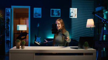 Neon ışıklı apartmandaki kız video bloglama kanalının hoş geldin girişini yakalamak için selfie çubuğuyla telefon kullanıyor. Oturma odasındaki neşeli şovmen, canlı yayında sadık seyircilerle bağlantı kuruyor, kamera B