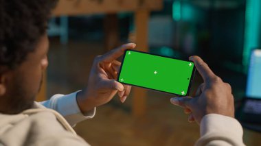 Afrikalı Amerikalı genç yetişkin, o koltukta dinlenirken oturma odasında aletlerini kullanarak yeşil ekrana bakıyor. Erkek kişi kendini rahat ve uzak işten memnun hisseder. Kamera A.