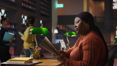 Kendine güvenen öğretmen notlarını okuyor ve kütüphane masasında öğretmenlik materyalleri hazırlıyor. Kendini pedagoji ve akıl hocalığına adadığını gösteriyor. Akademik destek için özel ders alıyorum. Kamera A.