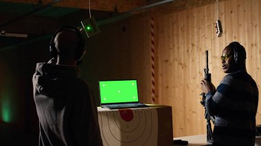Atış poligonundaki güvenlik görevlisinin yanındaki yeşil ekran dizüstü bilgisayar müşteriyi hedeflere ateş ederken izliyor, onu denetliyor. Model defter ve poligon uzmanı adama silah kullanmayı öğretiyor, kamera A