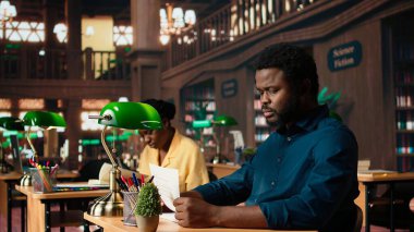 Siyahi üniversite öğrencisi endişeli ve öğrenme zorluğu çekiyor. İlgili bilgileri kütüphanedeki ders notlarından ezberlemeye çalışıyor. Erkek öğrenci sınavları geçmek için çalışıyor. Kamera A.