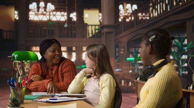 İlkokul öğrencileri bir profesörle matematik çalışıp akademik becerilerini kütüphane masasında geliştiriyorlar. Siyahi kadın müfredat materyallerini açıklıyor. Kamera A.