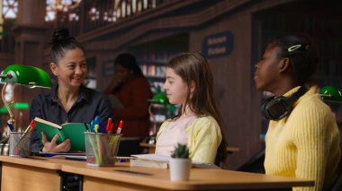 İlkokul öğrencileri, okul kütüphanesindeki çeşitli öğrenim ortamlarında ev ödevleri üzerinde çalışan bir öğretmenden destek alıyorlar. Profesör dersten önce öğrencilerine rehberlik ediyor. Kamera B.