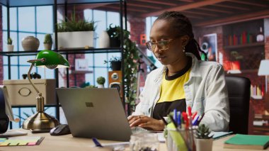 Evdeki Freelancer dizüstü bilgisayardaki analitik istatistiksel veri setlerini kontrol ediyor, evrak dosyalarıyla karşılaştırıyor. Not defterindeki anahtar performans göstergelerini belgelerle karşılaştırarak optimize eden kadın, kamera A