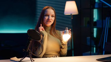 İçerik yaratıcısı stüdyoda ampulü sunuyor, profesyonel ekipmanlarla çekim yapıyor. Genç kız apartmanda pazarlama yapıyor, ışıklandırma ekipmanları ve kamera A 'yı sunuyor.