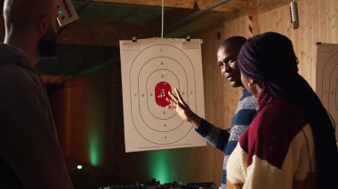 Galeri eğitmeni müşterilere atış puanı sistemini açıklıyor. Afrika kökenli Amerikan poligon uzmanları müşterilerine hedef tahtası sistemini öğretiyor, endüstri yönetmeliklerini tartışıyorlar, kamera A