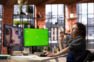 Kadın grafik tasarımcısı iyi organize edilmiş bir çalışma alanında fotoğraf rötuşlaması için düzenleme yazılımı ve yeşil ekran kullanır. Genç editör, yaratıcı bir sanat kariyeri için gerekli dijital araçları kullanıyor.