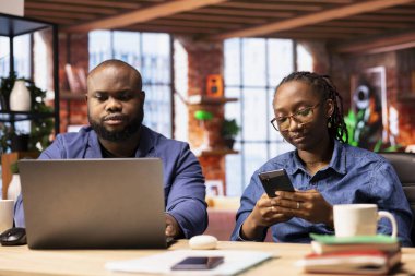 Siyahi bir çift masa başı işlerde uzaktan çalışıyor genç bir adam dizüstü bilgisayarda çalışıyor ve kadın müşterilerle akıllı telefondan iletişim kuruyor. Verimlilik için takım çalışması.