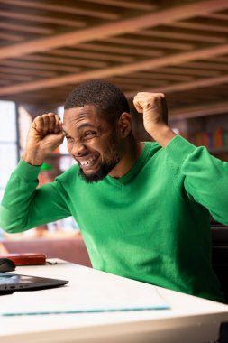 Neşeli, neşeli, Afro-Amerikalı Amerikalı adam evde kutlama yapıyor, olumlu duygular gösteriyor. Başarı, tezahürat ve el kol hareketi ile heyecanlanan BIPOC 'lu memnun kişi