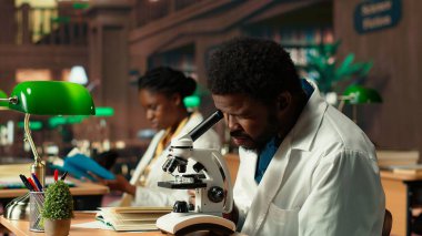 Mikroskop analizi sırasında virüs araştıran erkek öğrenci ve klasik bir kütüphanede akademik okuma. Tıp fakültesi öğrencisi biyolojiye ve sınav hazırlıklarına olan bağlılığını yansıtıyor. Kamera B.