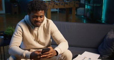 Kanepede oturmuş, sosyal medya ve mesajlarla eğlenen Afrikalı Amerikalı bir adam. Uzaktan çalışma molaları, dijital iletişim ve evde rahatlama. Kamera B.