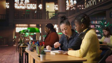 Kütüphane masasında ilkokul kızına rehberlik eden beyaz bir öğretmen, akademik gelişimlerine yardımcı olmak için çalışma kitapları ve notlarla özel dersler veriyor. Öğretmen akademisyenle konuşuyor. Kamera A.