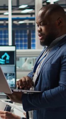 Fabrikadaki gülümseyen mühendisin dikey video portresi parametreleri kontrol etmek ve güneş panelinin dayanıklılığını artırmak için dizüstü bilgisayar kullanır. Mutlu Afrikalı Amerikalı araştırmacı verimli sürdürülebilir enerji geliştiriyor