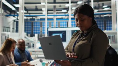 Happy solar panel tesis mühendisi verimliliği arttırmak için dizüstü bilgisayar kullanıyor. Gülümseyen Afrikalı Amerikalı kadın fotovoltaik fabrikası araştırma ve geliştirme bölümünde çalışıyor, not defteri, kamera A kullanarak.