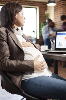 Müstakbel annenin masa başında oturup dizüstü bilgisayardaki şirket verilerine bakması. Hamile iş kadını dijital cihaz kullanıyor ve karnını nazikçe tuğla duvar odasında tutuyor..