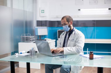 Tıbbi kayıtları incelemek ve hastane odasındaki e-postaları kontrol etmek için dizüstü bilgisayar kullanan erkek bir pratisyen. Maskeli doktor koruyucu ekranla masada oturuyor ve hasta randevularına hazırlanıyor..