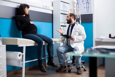 Dost canlısı erkek hekim, doğum öncesi ilerlemeyi açıklamak için dijital cihaz kullanarak beyaz hamile kadınla konsültasyon yapıyor. Doktor tableti tutuyor ve anne adayına hastane yatağında annelik tavsiyesi veriyor..