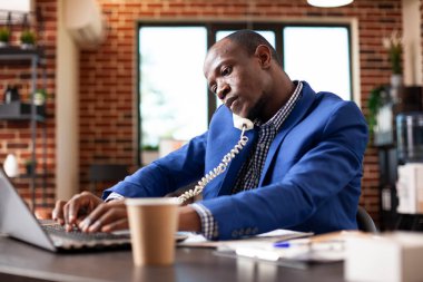 İşletme müdürü, ofis masasında bilgisayarla arama ve daktilo arasında birden fazla iş yapıyor. İş yerindeki girişimci, sabit hattan konuşuyor ve duvardaki kişisel bilgisayarındaki bilgileri güncelliyor..