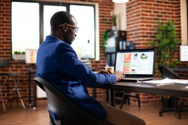 Erkek çalışan, şirket verilerine odaklanarak iş analizlerini başlangıç ofisinde dizüstü bilgisayarla analiz ediyor. Profesyonel siyahi adam dijital cihazdaki strateji planlarını gözden geçiriyor, tuğla duvar odasındaki masada oturuyor..