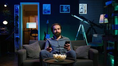 Oturma odasında televizyon izleyen, cep telefonundan arkadaşına mesaj atan, uzaktan kumandayı elinde tutan biri. Evde televizyon izleyen ve akıllı telefondan arkadaşla sohbet eden adam, kamera B