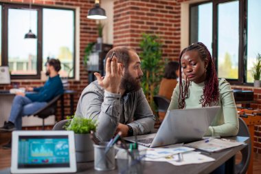 Beyaz adam ve siyah kadın birlikte finansal tabloları analiz ediyorlar dizüstü bilgisayar ve belgeler kullanarak, masalarda beyin fırtınası stratejileri kullanarak. Şirketin gelir artışı için işbirliği yapan ve stratejiler planlayan iş arkadaşları