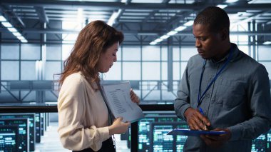 Bilgi işlem uzmanlarından oluşan bir ekip veri merkezi kontrol listesi evraklarını inceliyor. Afrikalı Amerikalı adam ve iş arkadaşı sunucu çiftliğinde işbirliği yapıyor. Belge dosyaları, kamera B