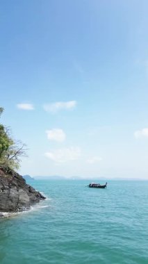 Sahil kayalıkları ve kumlu plajları olan tropikal bir ada kıyısı yakınındaki bir yelkenlinin dikey video görüntüsü çarpıcı bir perspektif, kristal berrak turkuaz okyanus suları ve egzotik kıyı şeridi olan bir tekneyi gösteriyor..