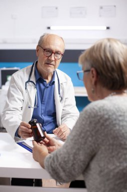 Yaşlı kadın hasta klinikte kıdemli erkek doktorla oturuyor ve iyileşme planı için reçeteli ilaç alıyor. Kafkasyalı yaşlı doktor, tıbbi muayeneden sonra emekli kadına ilaç sağlıyor..
