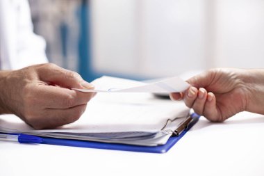 Kafkas sağlık hizmeti sağlayıcısı laboratuvar önlüklü hastaya reçeteli form sunuyor. Masanın üzerinde panosu olan tıbbi belgeleri tutan iki kişiye odaklanmış..