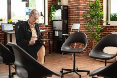 Elinde dosya olan son sınıf beyaz kadın modern odada tek başına oturuyor ve müşterilerle danışmanlık seansına hazırlanıyor. Gözlüklü yaşlı kadın terapist, duvardaki hasta notlarını inceliyor..