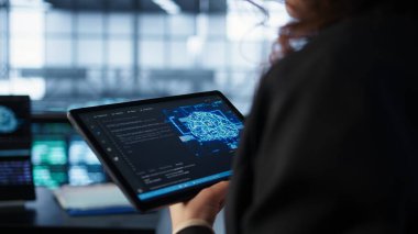 Veri merkezi teknisyeni savunmasızlığı önlemek için tablet üzerinde yapay zeka kullanıyor. Çalışan güvenlik duvarı kurallarını gözden geçirmek için aygıt kullanıyor, yapay zeka siber güvenlik protokollerini uyguluyor, kamera B kapatıyor.
