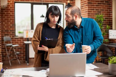 İş adamı proje toplantısında dizüstü bilgisayarı işaret ederken, bayan meslektaşı dikkatle dinliyor ve not alıyor. İş kadını elinde bir dosya tutuyor ve yeni açılan ofiste erkek iş arkadaşıyla tartışıyor..