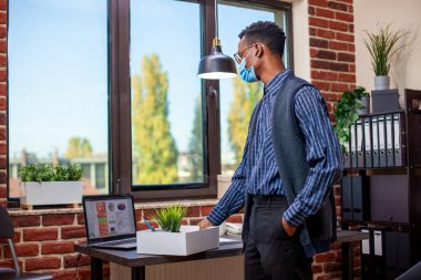 Masa başında maske takan bir işletme danışmanı bir kutu iş malzemesi ayarlıyor, saksı da dahil. Siyahi girişimci önemli proje detaylarını gözden geçiriyor ve ayrılmaya hazırlanıyor.