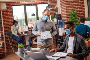 Beyaz adam, siyahi meslektaşı ile güvenli bir ofis ortamında yaptığı bir konuşma sırasında bir tabletle veri paylaşıyor. Erkek iş arkadaşları, her ikisi de tuğla duvar iş yerinde maske takarak bir masada iş birliği yapıyorlar..