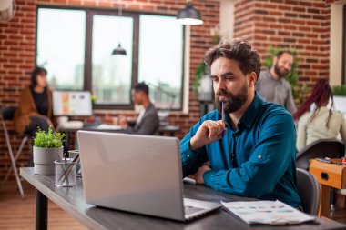 Erkek araştırmacı elinde kalemle belgeleri inceliyor, masadaki proje geliştirmeye odaklanmış. Kafkas çalışanı tuğla duvar ofisinde oturmuş, çenesini tıkırdatıyor ve iş planlarını dizüstü bilgisayardan inceliyor..