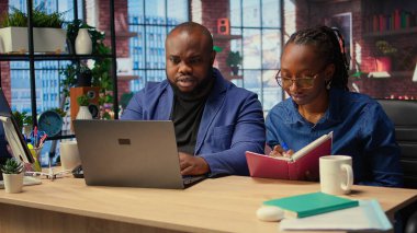 Defterdeki hesap çizelgelerini tamamlayan, hücrelere sayısal verileri giren, toplamları hesaplayan bir kadın. Afrikalı Amerikalı çift, bir muhasebe görevi için internet bilgilerini inceliyor. Kamera A.