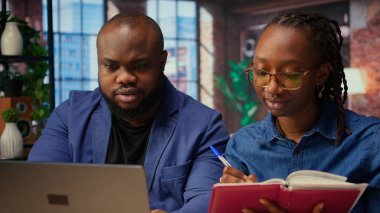 Siyah uzaktan kumandalı çalışanlar takvim randevularını dizüstü bilgisayar ve defter kullanarak düzenliyor, kağıtları kontrol ediyor ve önemli görevleri zamanında senkronize ediyor. Erkek ve kadın evdeki dosyaları gözden geçiriyorlar. Kamera A.