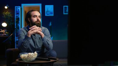 Televizyon ekranında gösterilen ilginç filmle kanepede oturan adam. Neon ışıklı oturma odasında canlı yayın yapan kişi, eğlenceli film sahnesinden büyülenmiş, dikkat çeken, kamera B