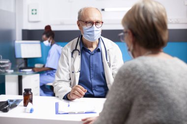 Kıdemli kadın ve yaşlı erkek doktor kontrol sırasında panodan alınan teşhisleri tartışıyorlardı. Yüz maskeli emekli doktor klinikteki kadın hastaya tıbbi tavsiyelerde bulunuyor..