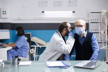 Genç doktor, klinikte oturan yaşlı hasta bir adam üzerinde otoskop kullanıyor ve sağlık muayenesi sırasında doğru teşhisi koyuyor. Erkek otolog, tıbbi aletle emekli hastaya kulak muayenesi yapıyor..