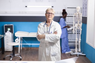 Laboratuvar önlüğü giyen kıdemli kadın doktor klinikte duruyor. Afrikalı Amerikalı hemşire arka planda kabineyi düzenliyor. Tıbbi odada kollarını kavuşturmuş kameraya bakan yaşlı bir doktor.
