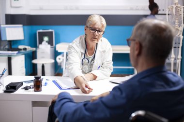 Beyaz yaşlı adam, kadın hekimin sağlık tedavisini açıklamasını dinliyor. Emekli erkek tekerlekli sandalye kullanıcısı yaşlı kadın doktoruna iyileşme süreciyle ilgili bilgi veriyor..