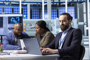 Üretim fabrikası uzmanı dizüstü bilgisayarla sürdürülebilir üretim araştırma ve geliştirme verilerini analiz ediyor. Yatırımcılar yatırım stratejilerinin karlılığını değerlendirmek için defter üzerindeki belgeleri gözden geçirirler