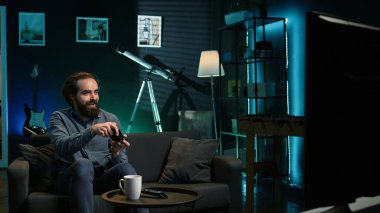 Neon ışıklı evde video oyunu oynayan mutlu bir adam, elinde kontrol cihazı tutuyor. Oyuncu çevrimiçi turnuvaya katılmak için heyecanlı, profesyonel joypad kullanarak diğer oyuncularla mücadele ediyor